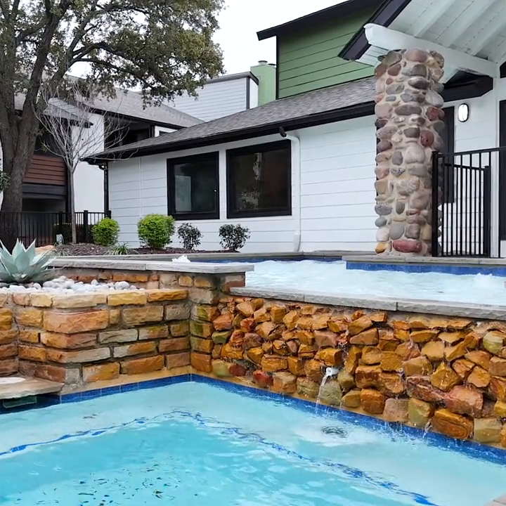 A serene backyard scene featuring a modern swimming pool surrounded by a stone wall. The pool is bordered by lush landscaping with agave plants and small shrubs, creating a tranquil atmosphere. The house in the background has a contemporary design with large windows and a covered patio area, enhancing the luxurious feel of the outdoor space. The overall setting suggests a peaceful retreat perfect for relaxation and leisure.